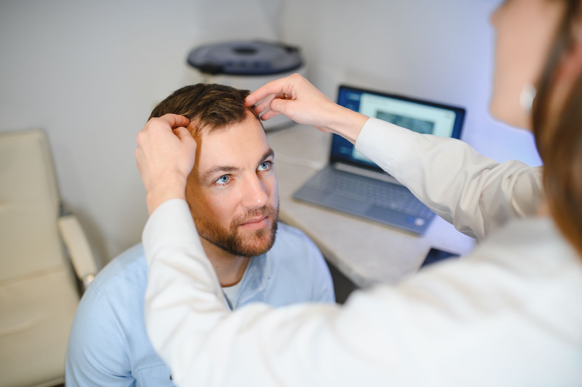 Doctor examining a patient’s forehead in a clinic office, with a laptop in the background