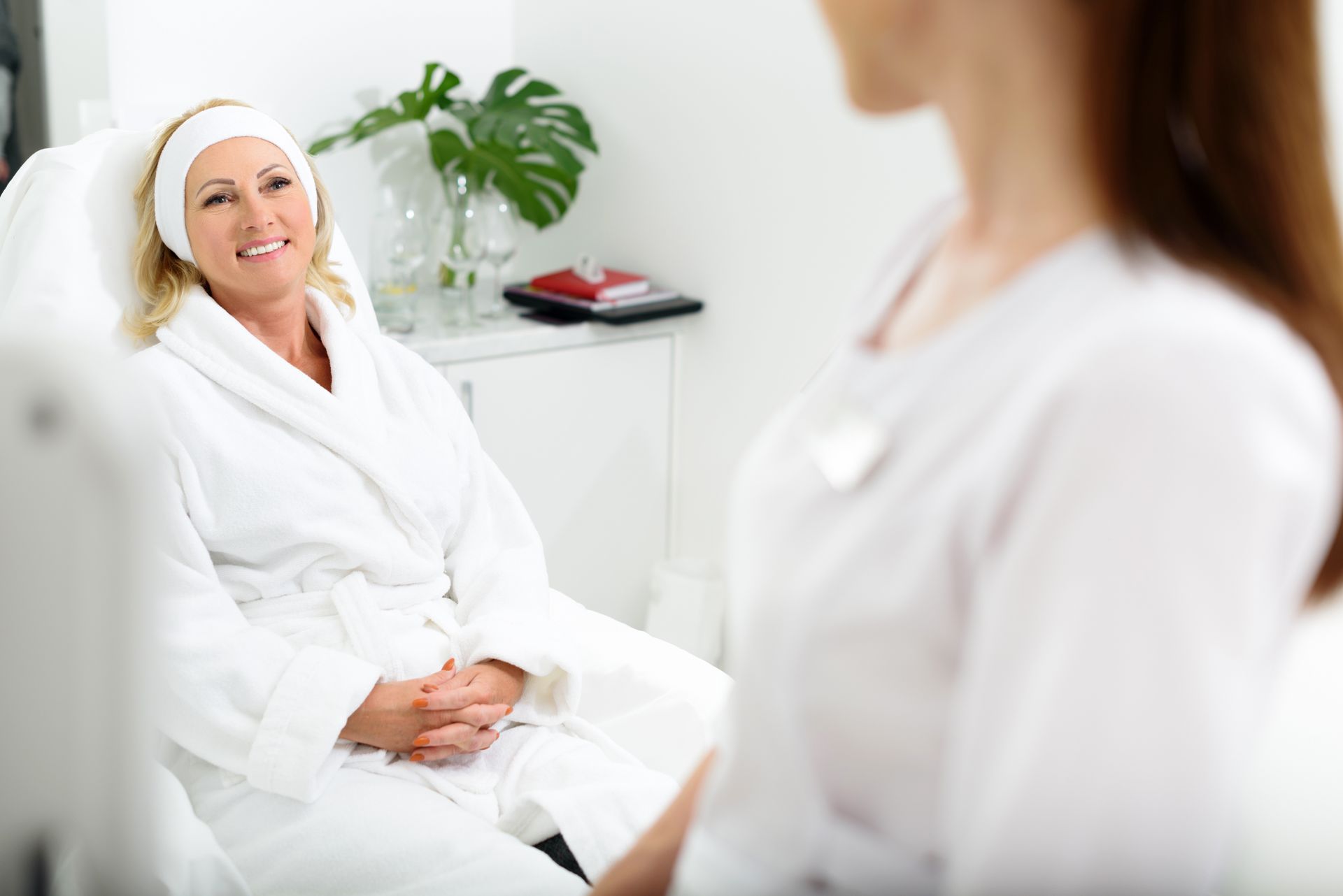 Woman in a white robe smiling while seated in a bright clinical room, speaking with another person.