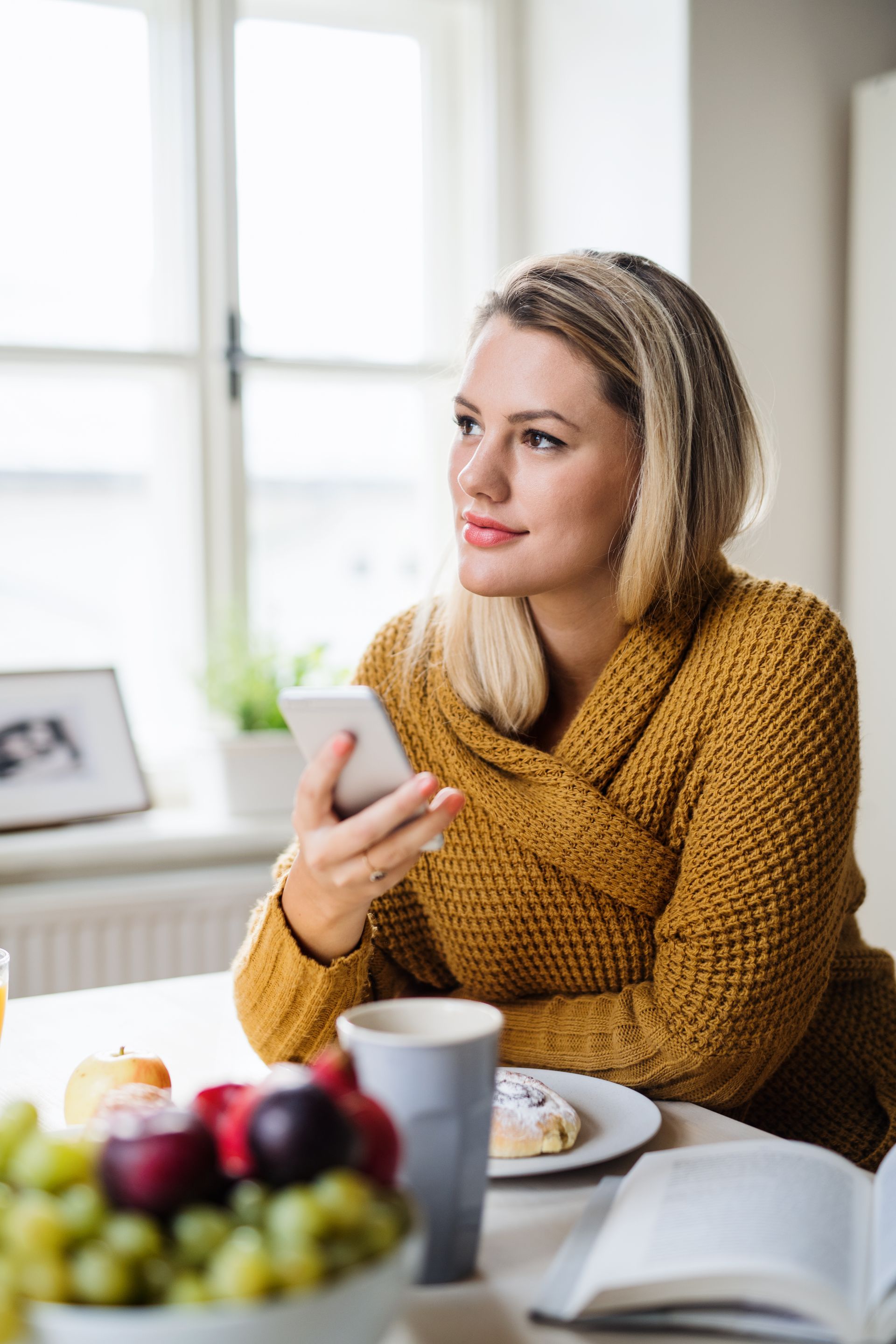 Woman in a mustard sweater holding a phone at a table with coffee and fruit by a bright window