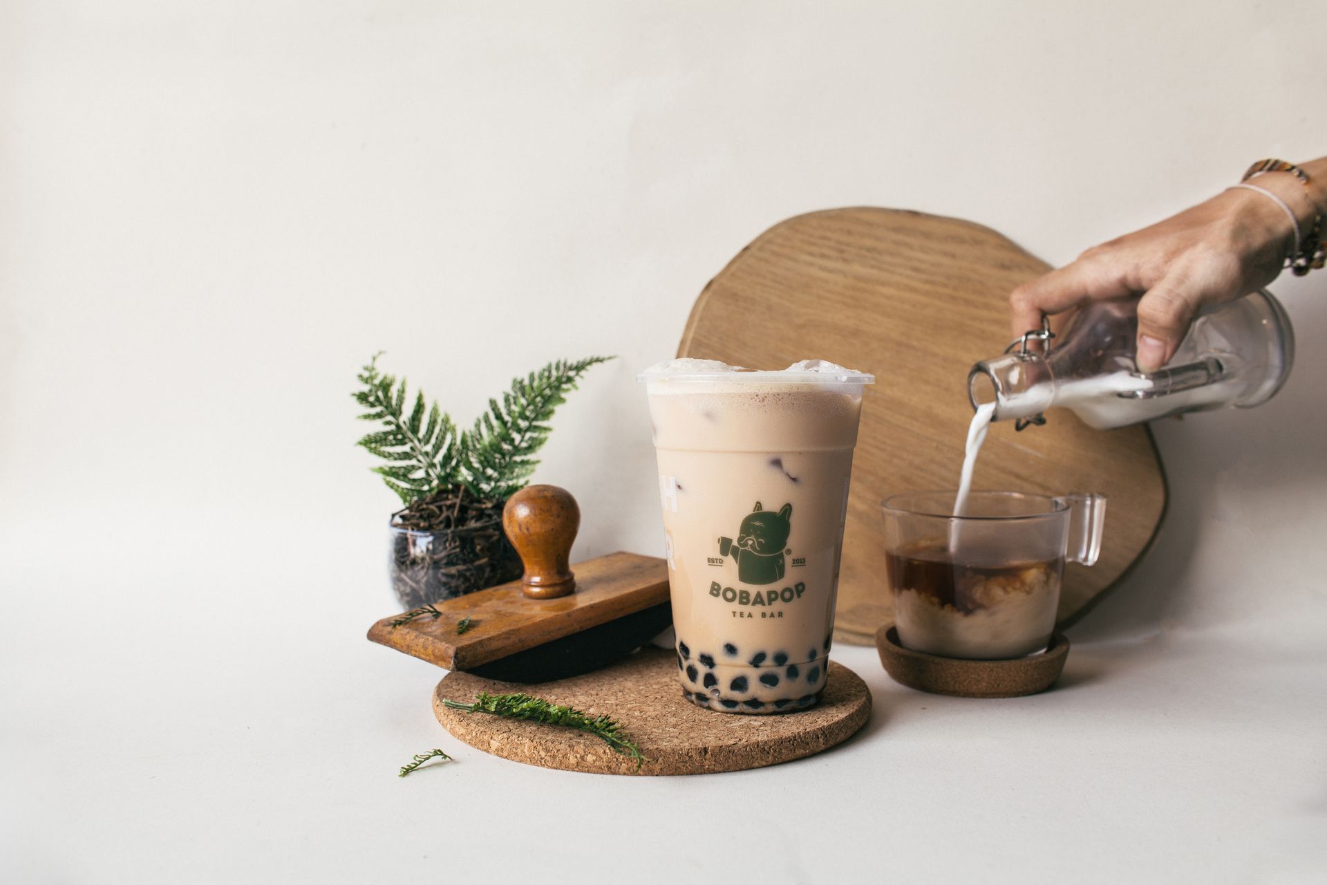Hand pouring iced coffee into a glass beside a clear bubble tea cup, fern, and wooden tray
