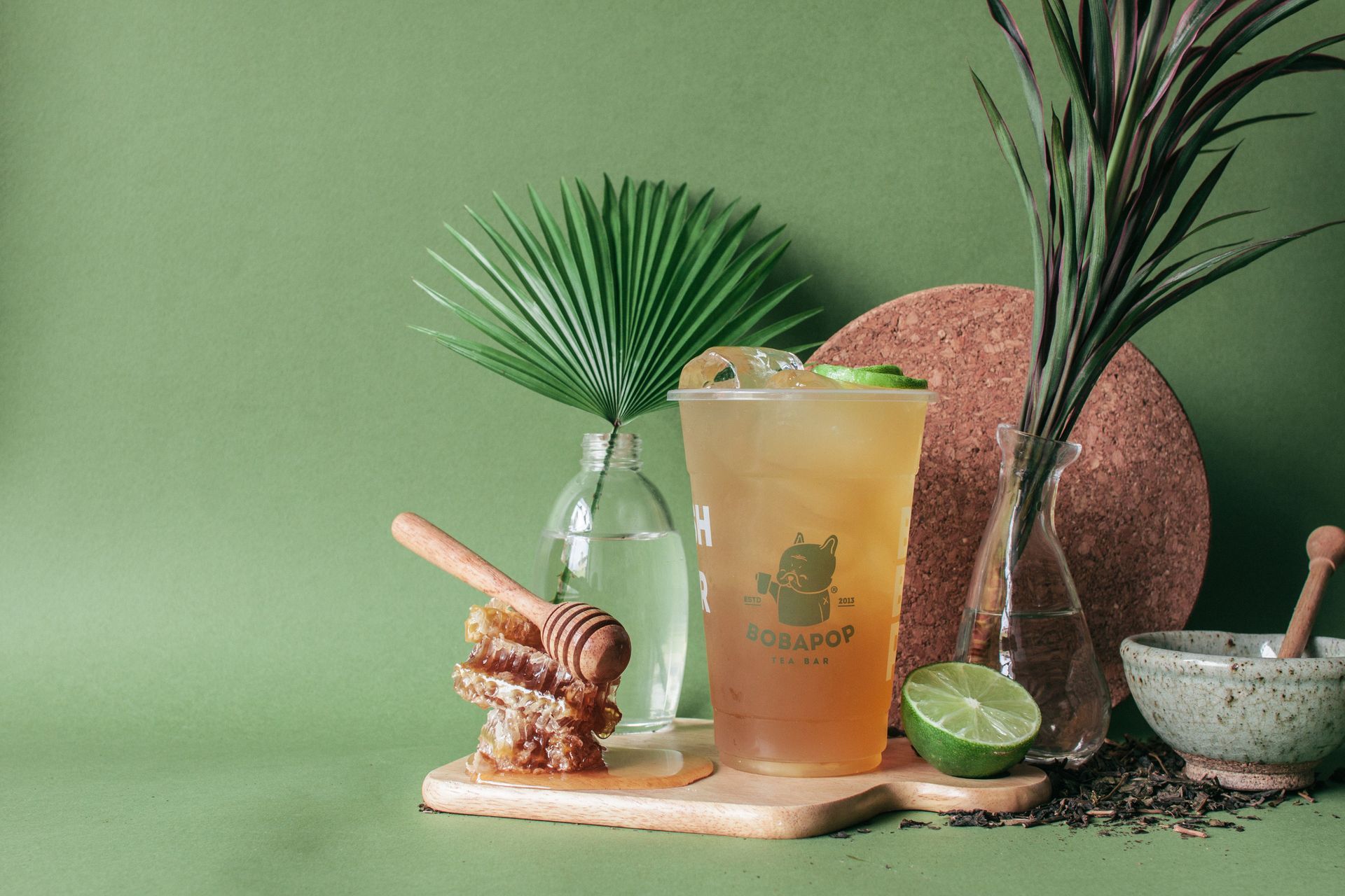 Green cocktail with lime on a tray beside a bottle, fan leaf, and mortar on a mint-green background