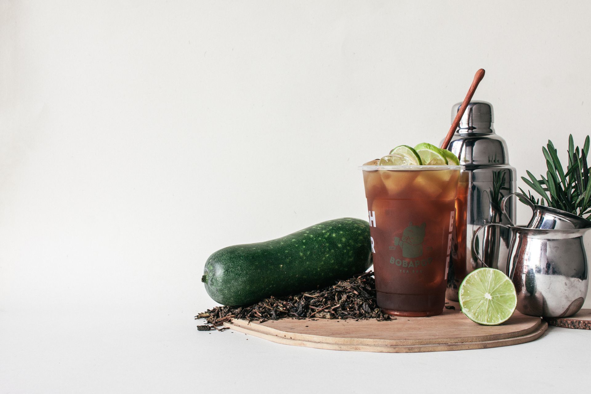 Cucumber, lime, and iced cocktail in a glass on a wooden board with bar tools and herbs