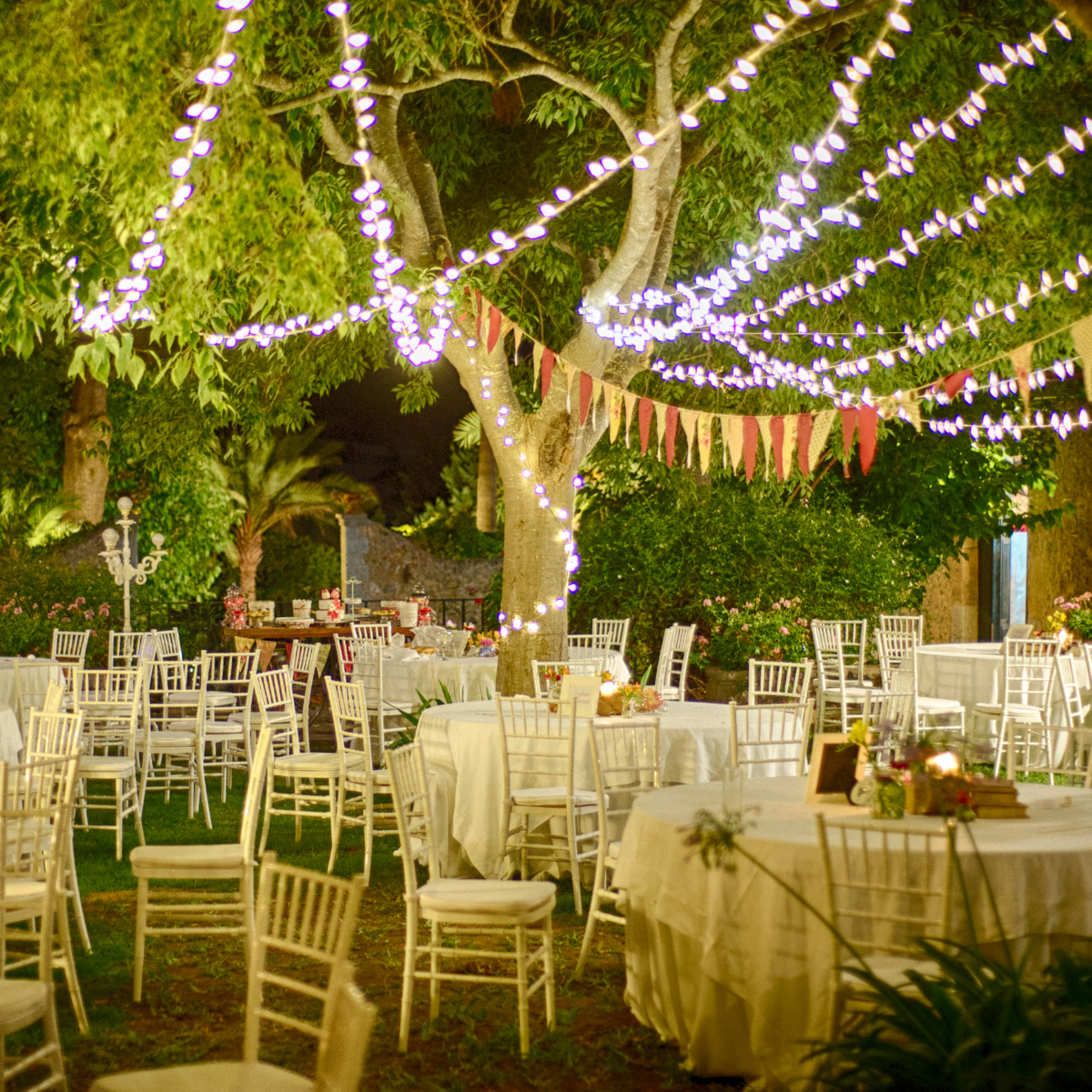 Outdoor evening wedding reception with string lights, white chairs, and round tables under trees