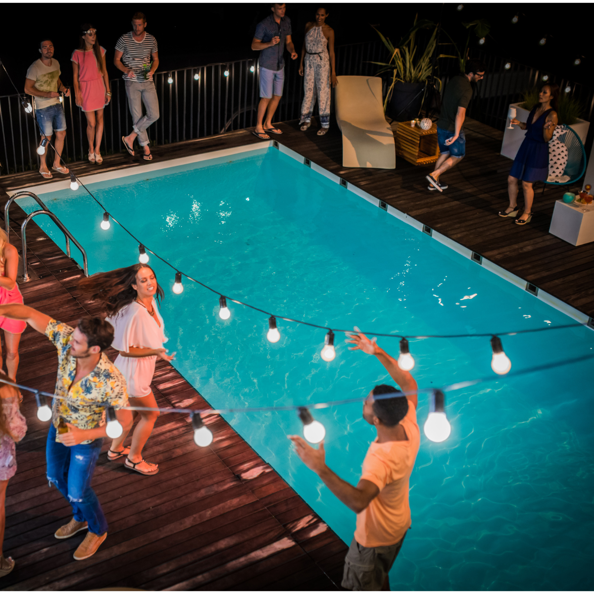 People gathered around a lit pool at night, dancing on a wooden deck with string lights.