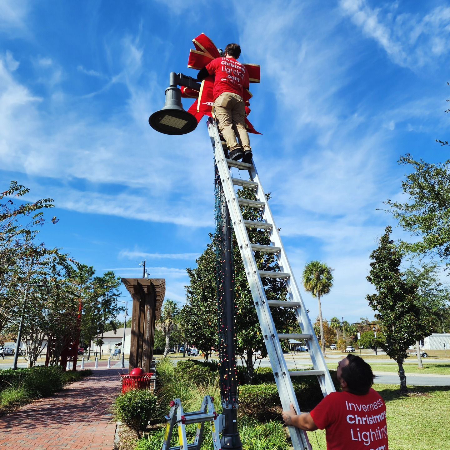 Person in red jacket climbs a ladder to install a streetlight in a park under a blue sky.