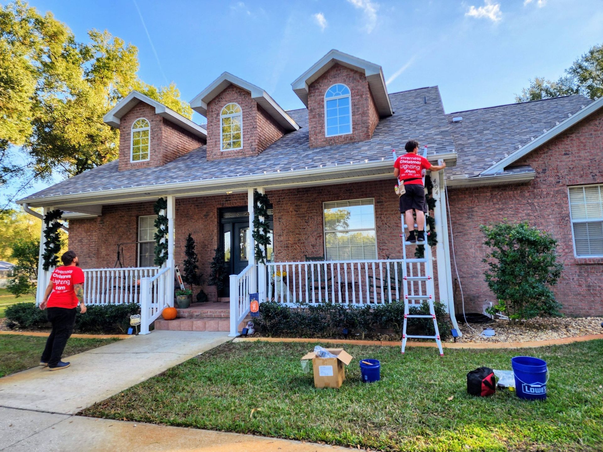 Three people decorating a brick house with Christmas lights, one on a ladder and two on the lawn.