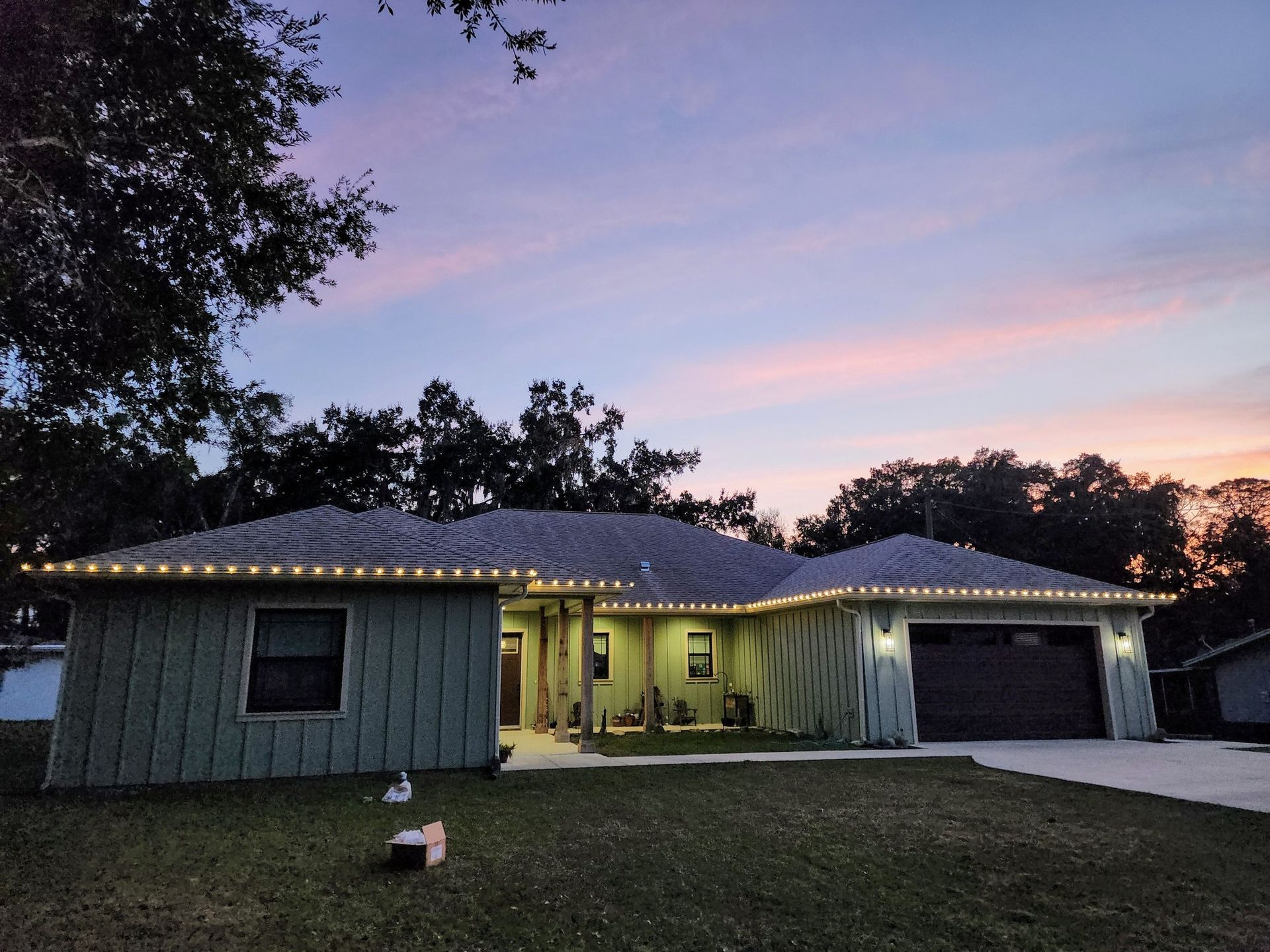 Single-story house at dusk with warm porch lights, a lit roofline, and a driveway under a pink sky.