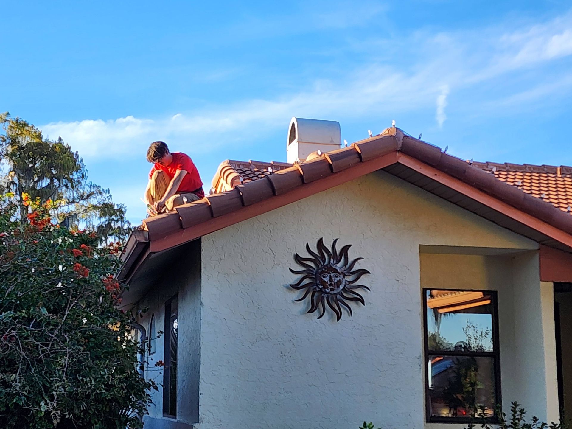 Worker on a house roof beside a chimney, wearing a red shirt under a blue sky.
