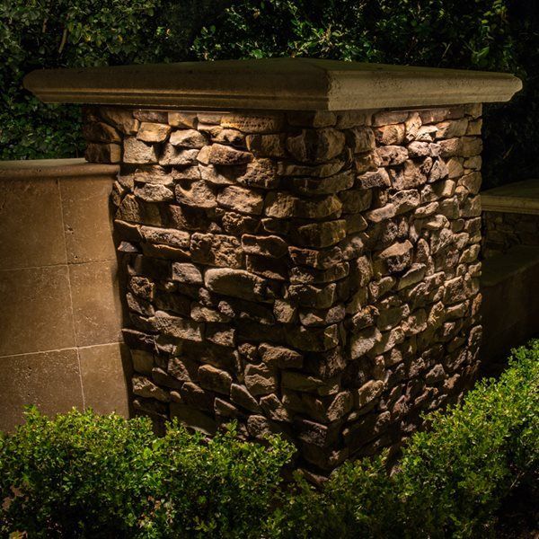 Stone column with a tan cap in a garden at night, surrounded by green shrubs and dim lighting