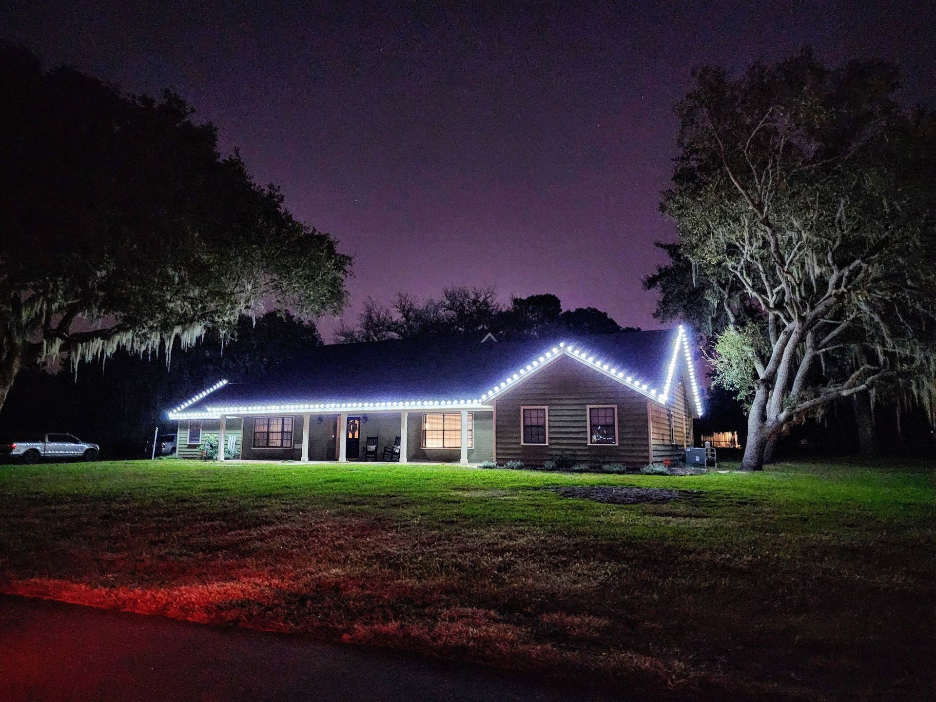 House outlined in white Christmas lights at night, with a dark lawn and trees under a purple sky