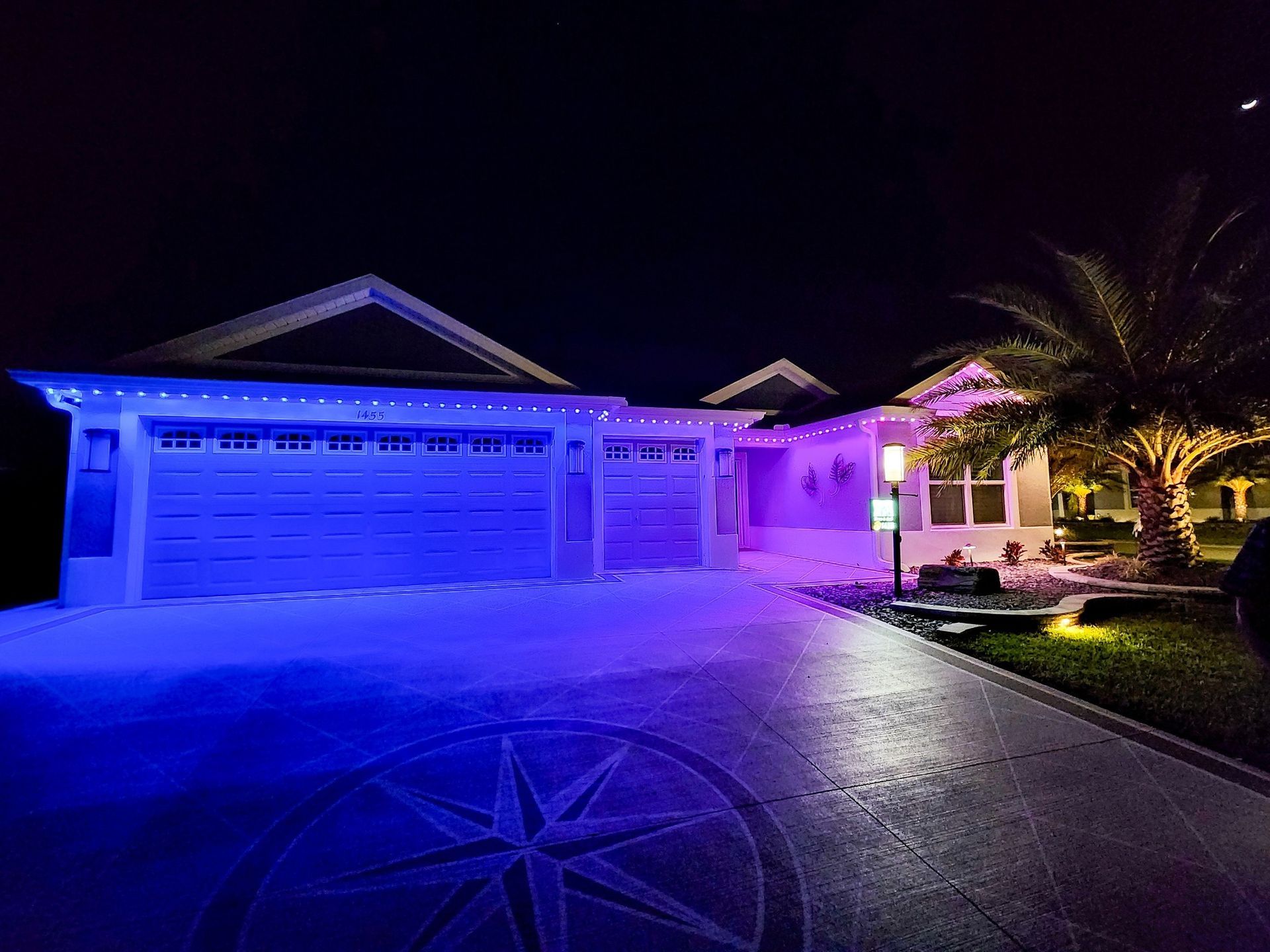 Blue and purple-lit suburban house at night with a two-car garage and palm tree in front