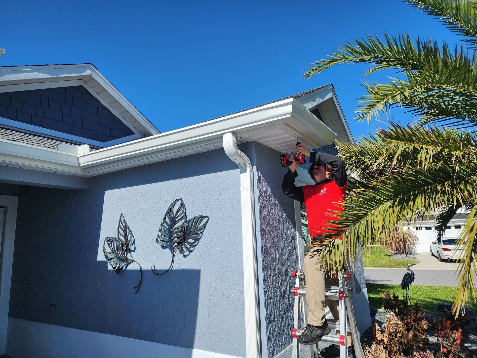 Worker on a ladder cleaning a house’s white gutter under a blue sky