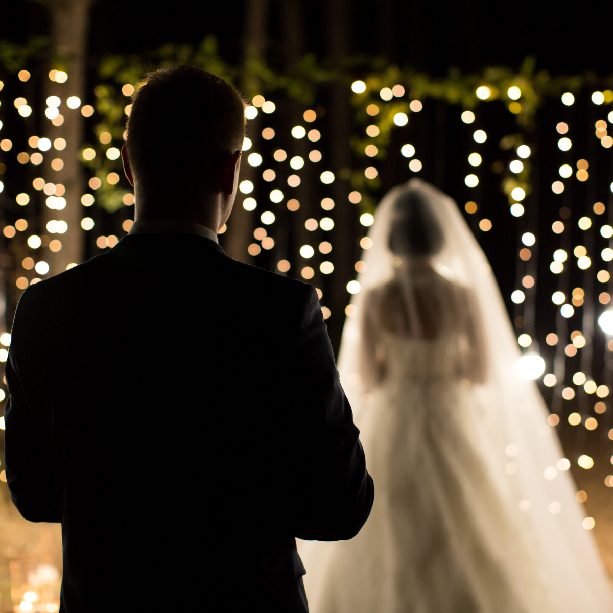 Silhouetted man watching a bride in a white veil before glowing string lights at a wedding