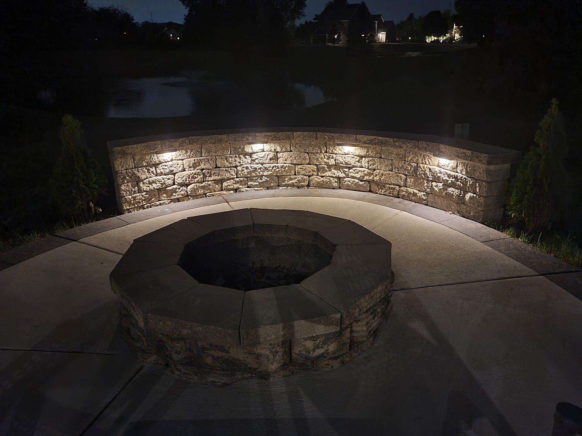 Stone fire pit in a landscaped backyard patio at dusk, lit by warm outdoor lights.