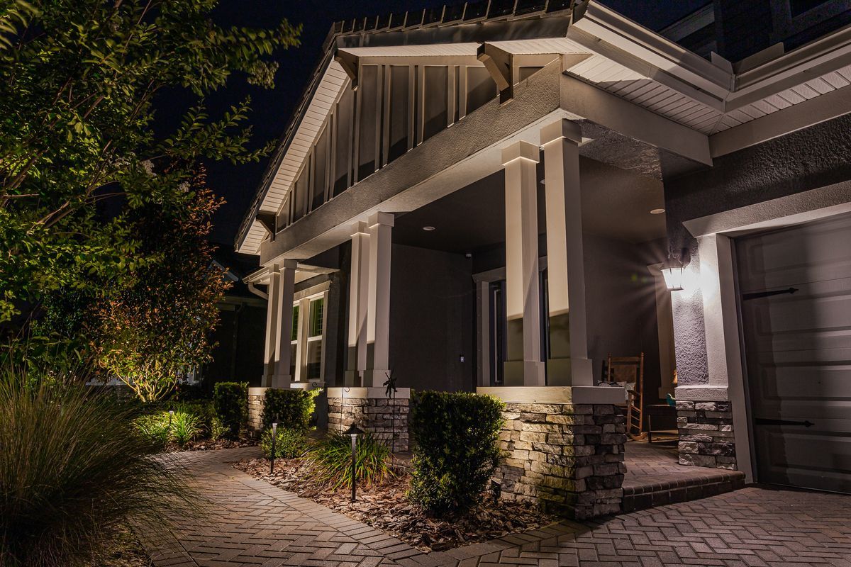 Nighttime view of a well-lit stone house with a porch, gabled roof, and trees beside a paved driveway.