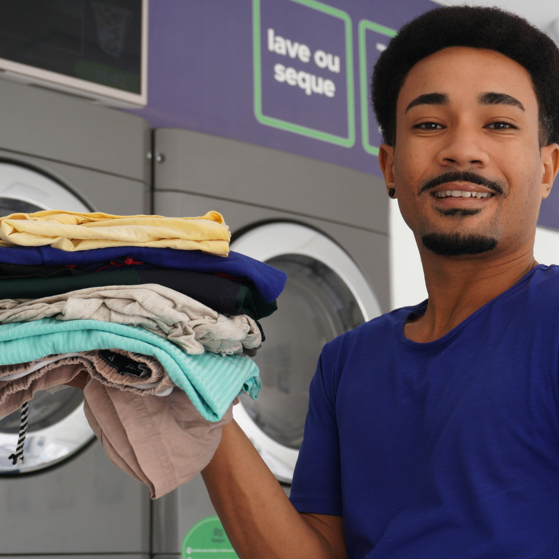 Person holding folded laundry in a laundromat, smiling beside stacked clothes and washing machines.