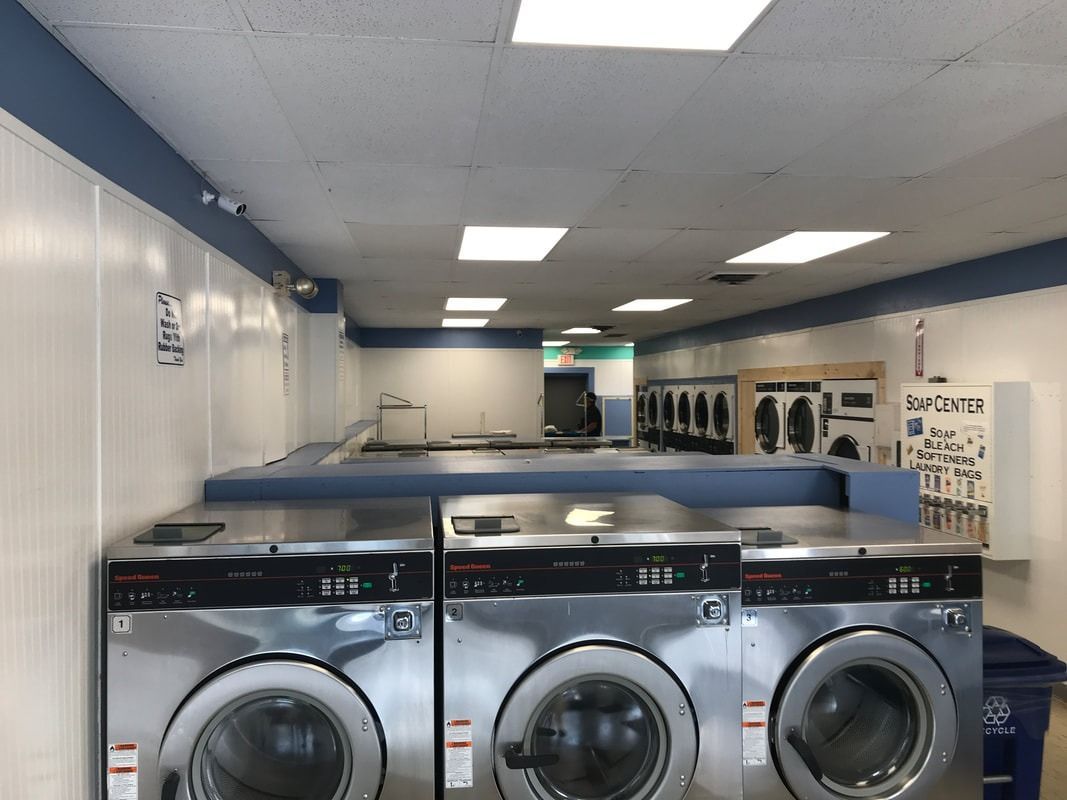 Laundry room with rows of front-load washers and blue folding tables under bright ceiling lights