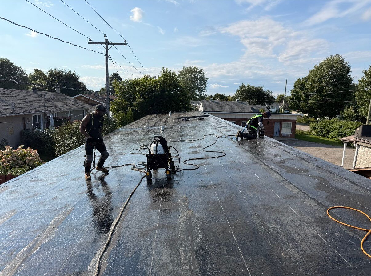 Des ouvriers appliquent un revêtement de toiture noir sur un toit plat à l’aide d’un tuyau et d’outils, avec des maisons et des arbres en arrière-plan