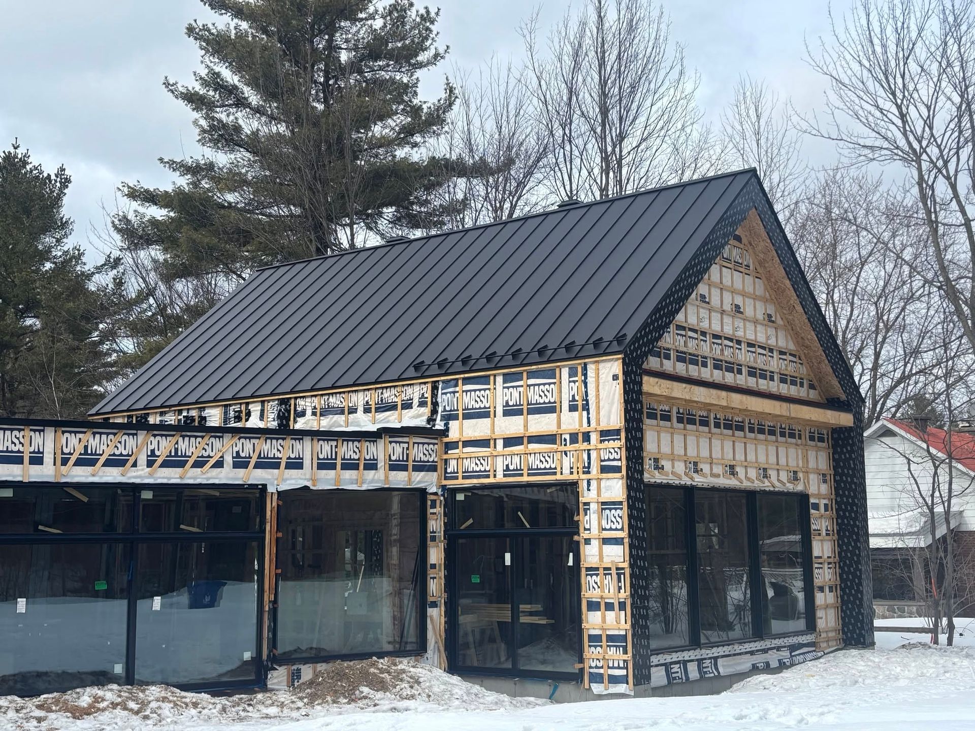 B&acirc;timent en construction avec toit en t&ocirc;le m&eacute;tallique gris et fa&ccedil;ade en bois partiellement recouverte de panneaux isolants.