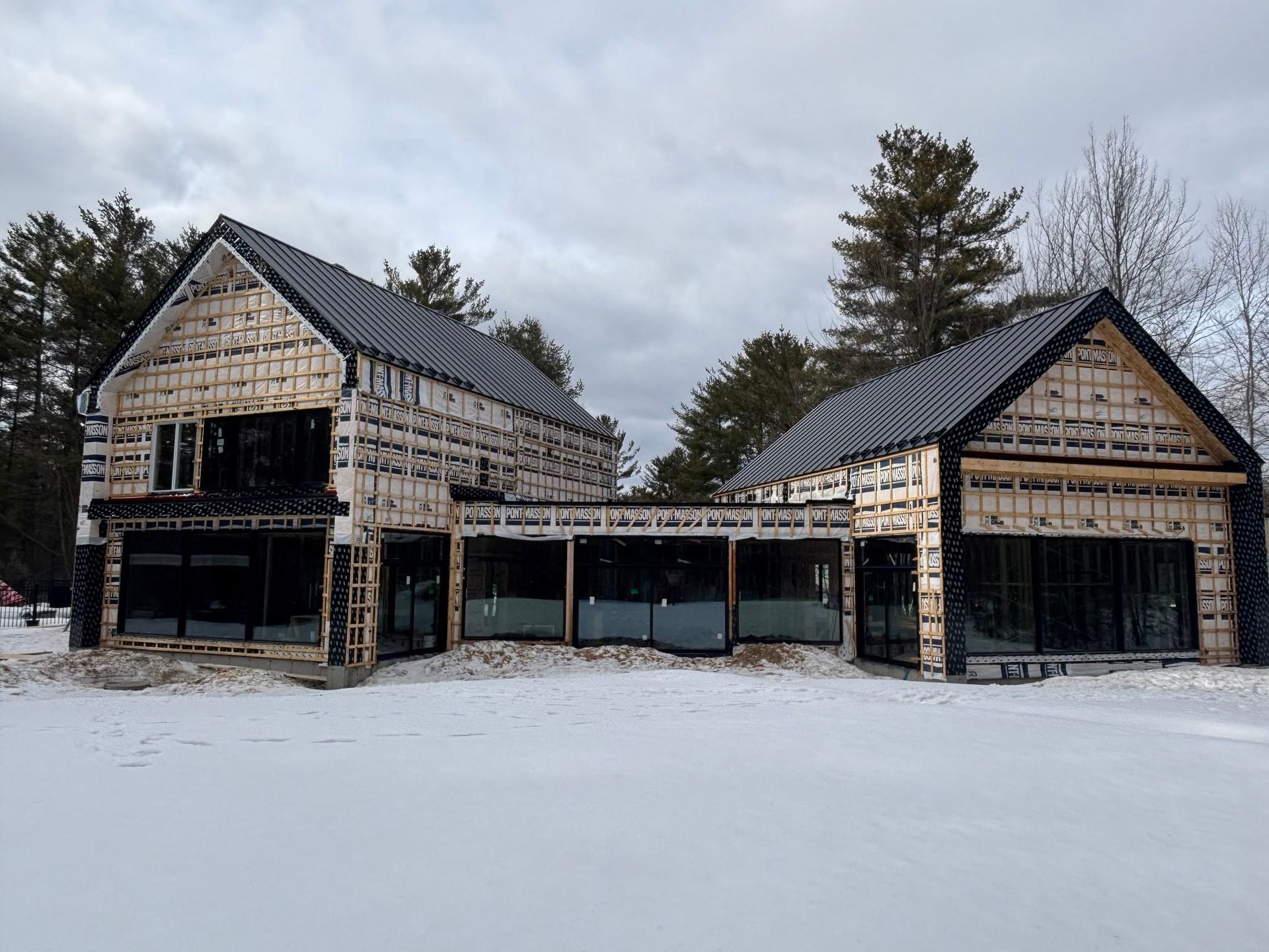 Maison inachevée couverte de neige, avec un toit sombre et une ossature en bois, sous un ciel nuageux