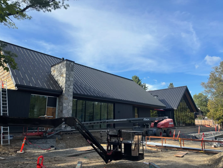 Maison moderne en construction avec un toit noir, une cheminée en pierre et du matériel de chantier à l’avant