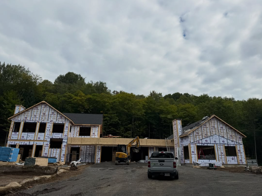 Maison en construction avec sa charpente apparente, un camion devant et des piles de bois sous un ciel nuageux