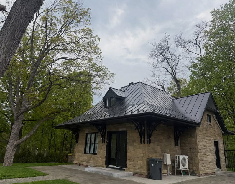 Petit bâtiment en pierre avec un toit en métal sombre, entouré d’arbres sous un ciel nuageux.