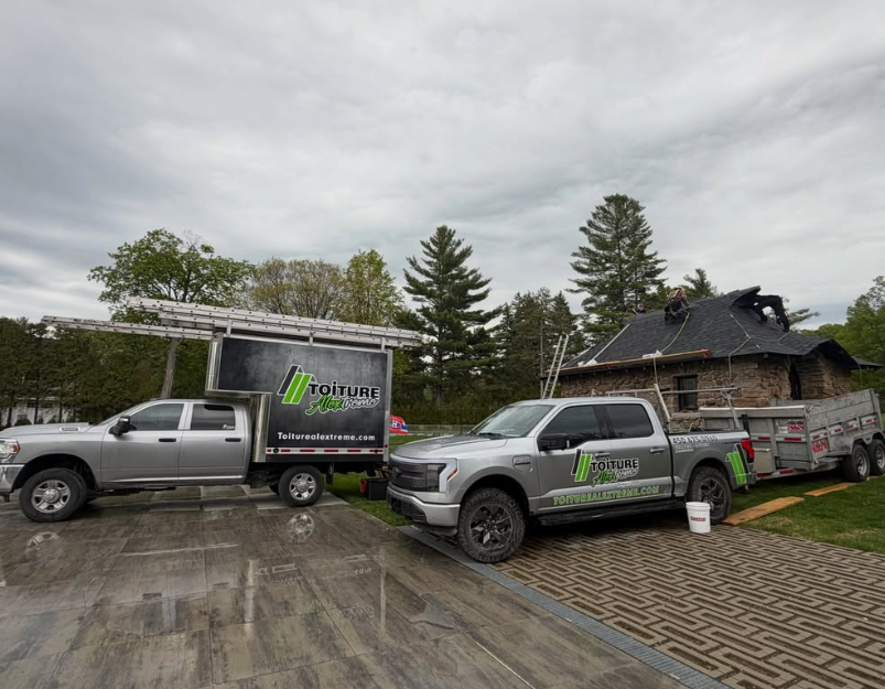 Deux camionnettes argentées avec des remorques de travail sont garées dans une allée mouillée près d’une maison et d’arbres, sous un ciel nuageux