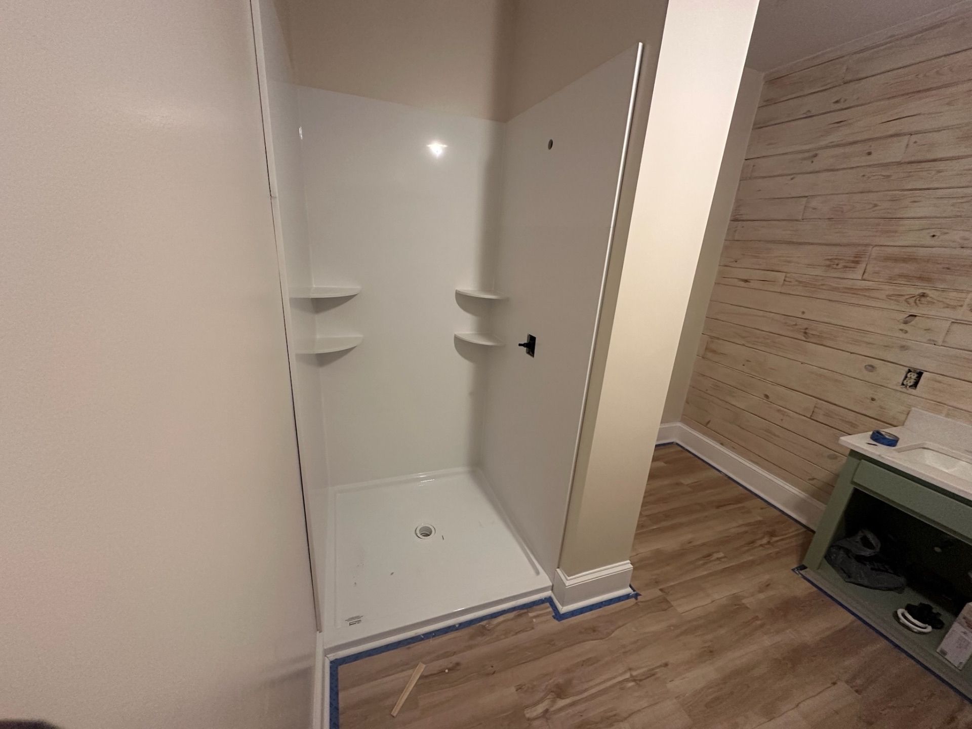 Empty walk-in shower with white tile, built-in corner shelves, and wooden flooring beside it.
