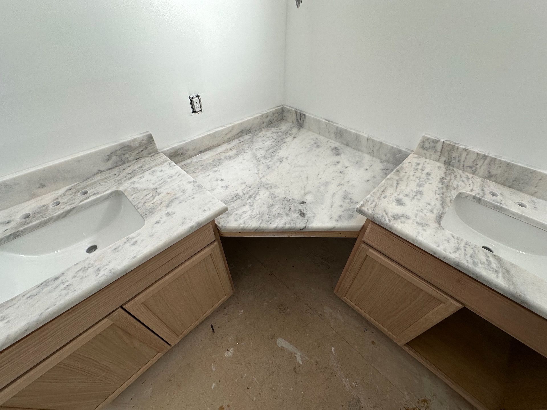 Corner bathroom vanity with two sinks and white marble countertop, showing open cabinet spaces below.