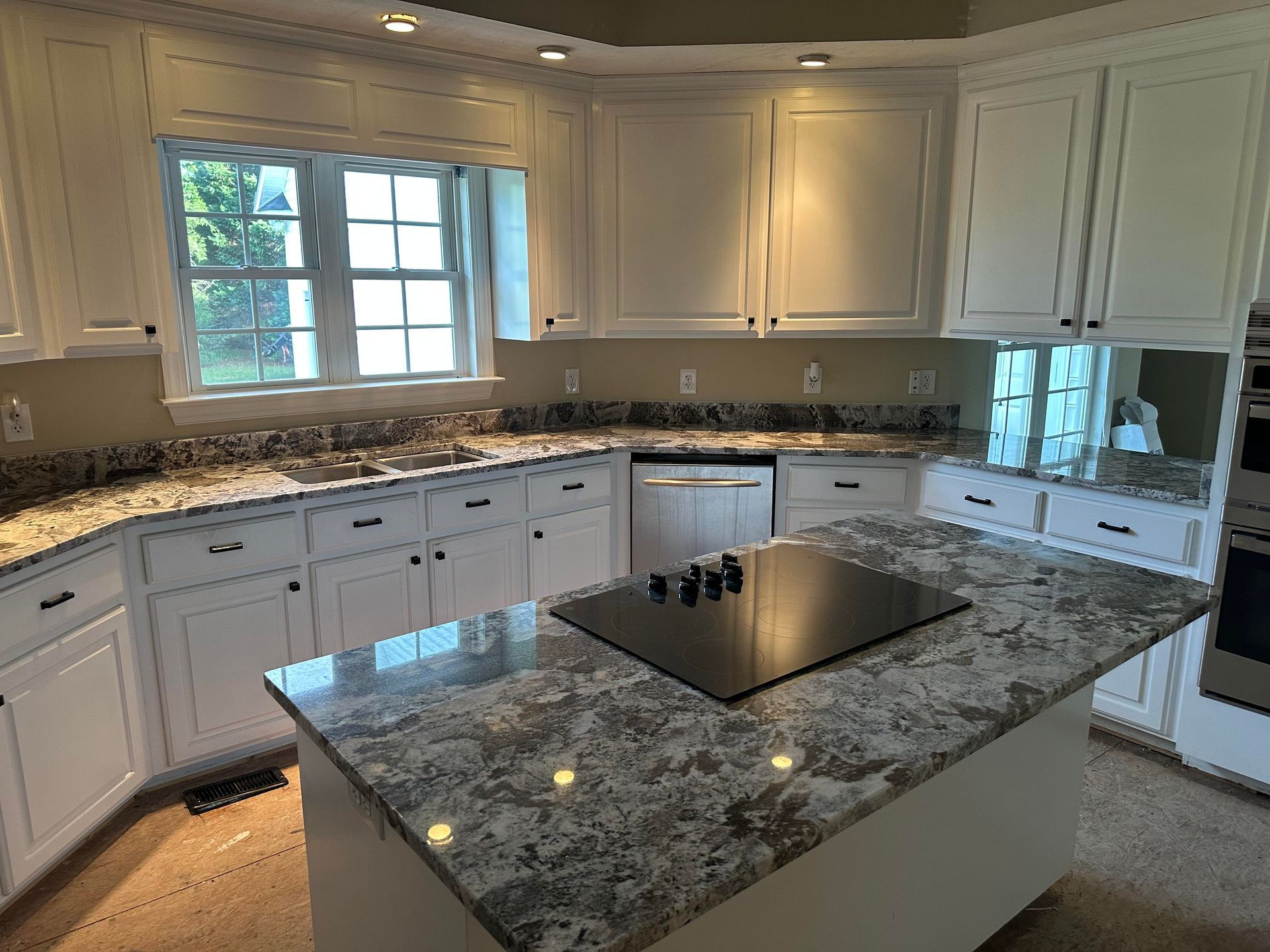 White kitchen with granite countertops, island with cooktop, cabinets, and a window.