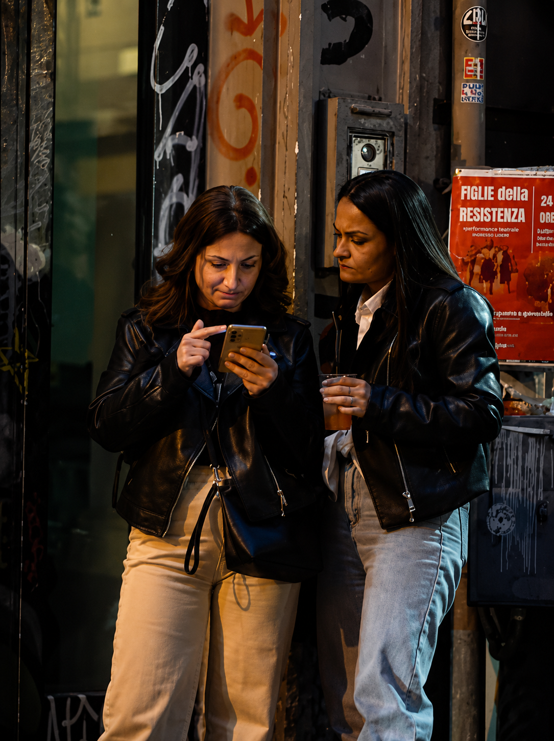 Due donne con indosso giacche nere guardano un telefono su un marciapiede cittadino, accanto a un muro ricoperto di graffiti.