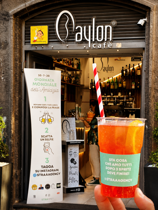 Cocktail in bicchiere trasparente con bevanda rosso-arancione all'esterno di un bar con menù e bottiglie all'interno.