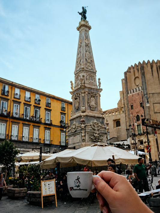 Una mano che regge una tazza di caffè in una piazza affollata con un alto monumento decorato e ombrelloni da caffè.