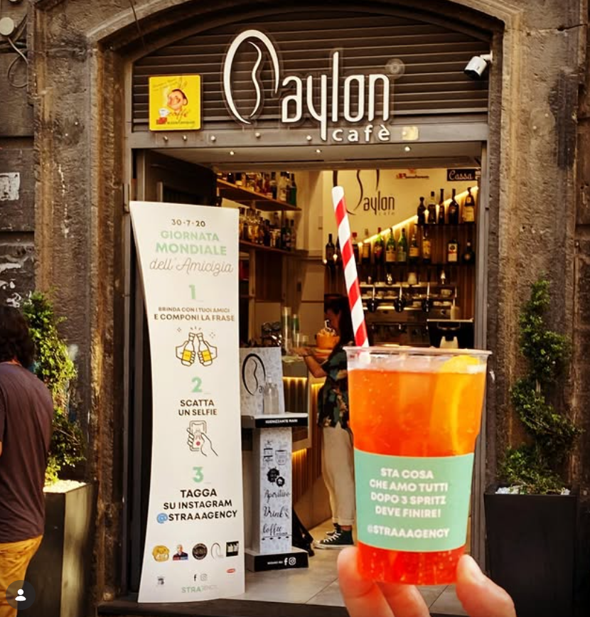 Vetrina di una caffetteria con una grande bevanda colorata in primo piano e un cartello con il menù all'esterno.