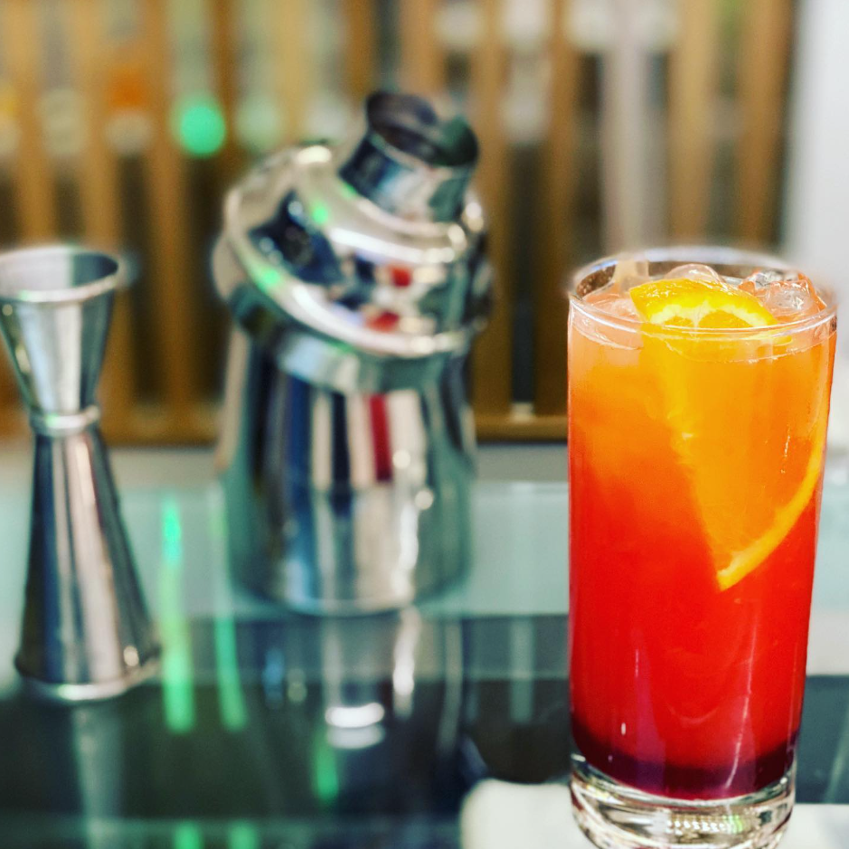 Cocktail rosso-arancio con una fetta d'arancia su un bancone da bar, accanto a uno shaker e un misurino in metallo.