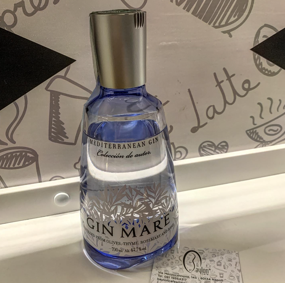 Bottiglia blu di Gin Mare con tappo argentato su un tavolo in stile caffetteria, con uno sfondo a tema latte macchiato.