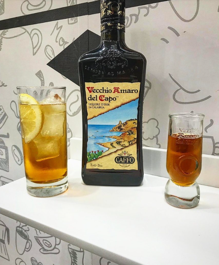 Una bottiglia di liquore Vecchio Romagna del Capo tra due bicchieri di bevanda ambrata con ghiaccio su uno scaffale bianco.