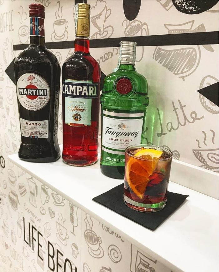 Tre bottiglie di cocktail su uno scaffale bianco, con una bevanda rosso-arancio in un bicchiere basso su un sottobicchiere nero.