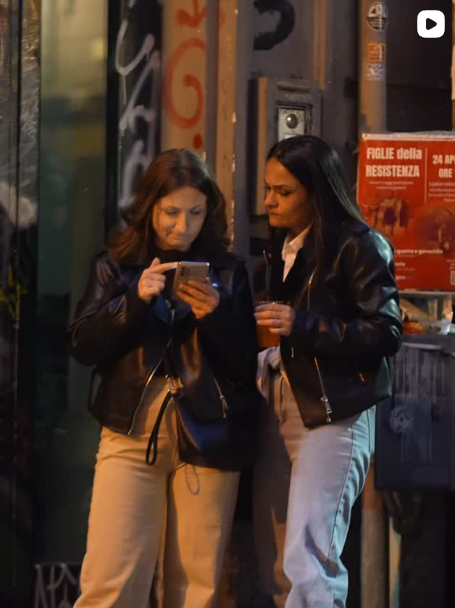 Di notte, due persone con indosso giacche scure sono in piedi all'aperto e guardano un telefono davanti alla vetrina illuminata di un negozio.