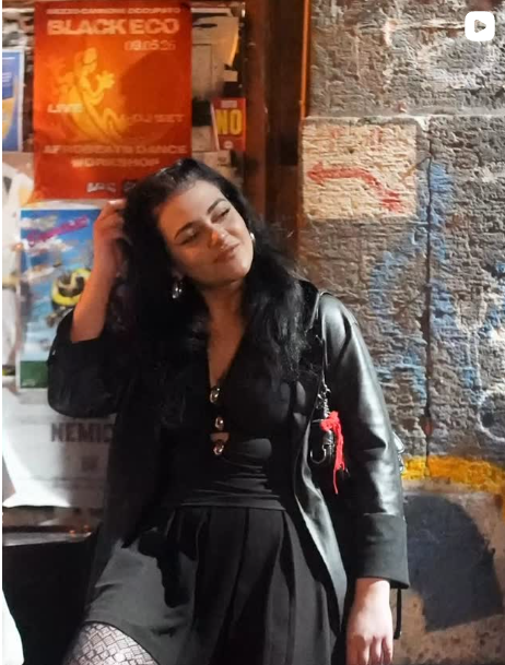 Una donna vestita di nero è appoggiata a un muro ricoperto di poster e si tocca i capelli in un contesto urbano.