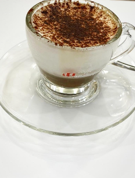 Una tazza di cappuccino in vetro con cacao in polvere è appoggiata su un piattino trasparente.