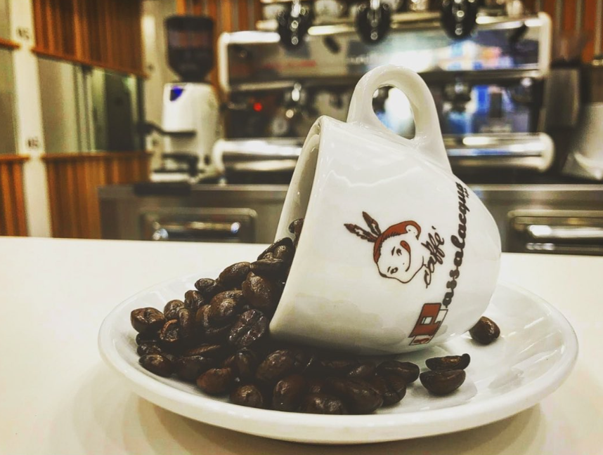 Tazza bianca rovesciata su un piattino, con chicchi di caffè che si rovesciano su un tavolo, sullo sfondo sfocato di un bar.