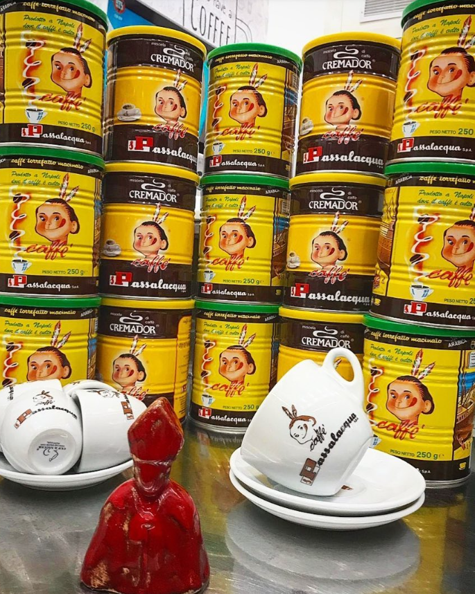 Lattine gialle impilate con una faccina stilizzata, affiancate da due tazze da tè bianche e una statuetta rossa su uno scaffale.