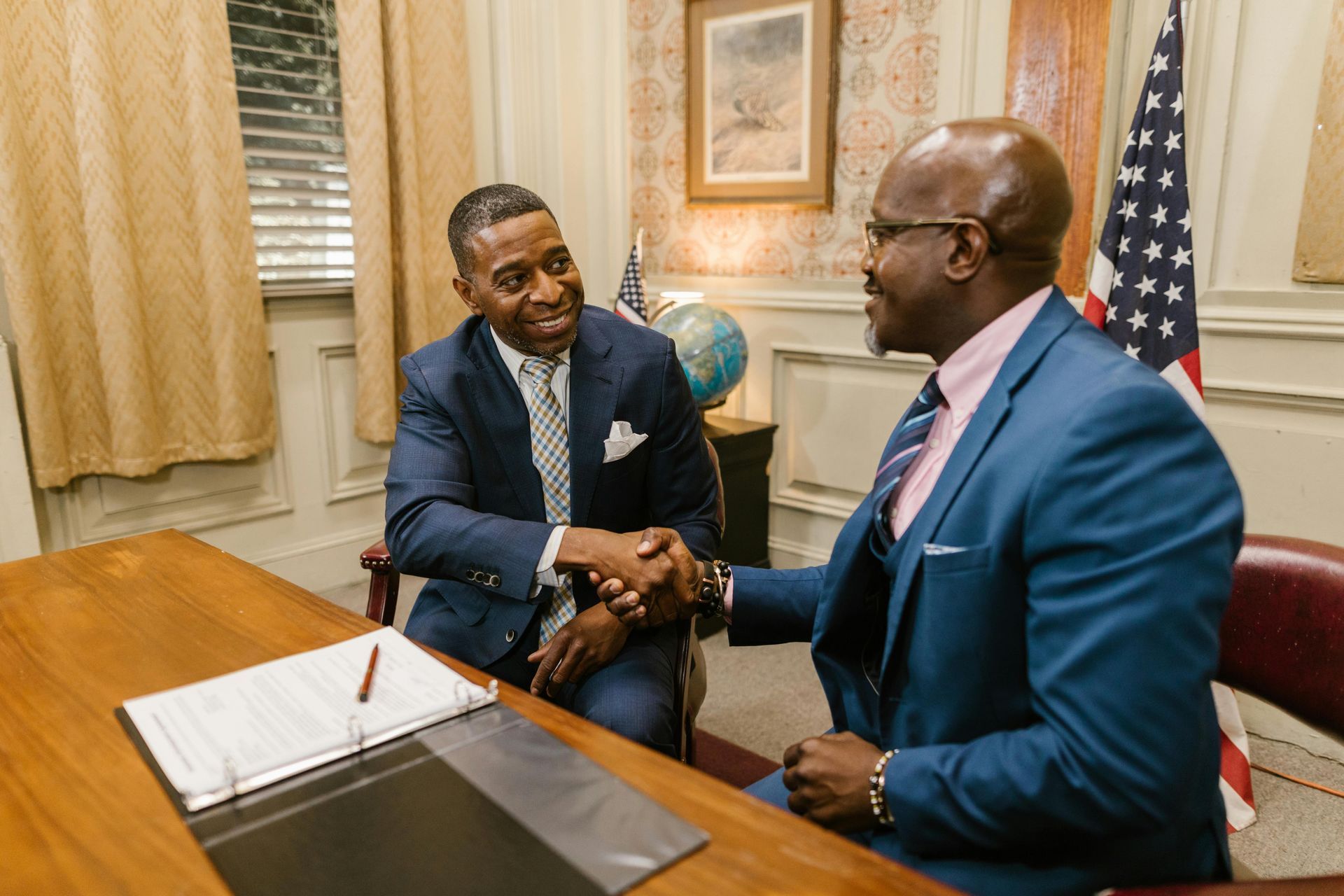 Real photo of two professional black men sitting in an office shaking hands with an American flag behind them.