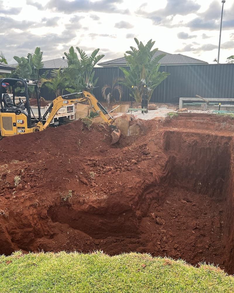 A Yellow Excavator is Digging a Hole in the Dirt in Front of a House — Norcoast Diggers in Lismore, NSW