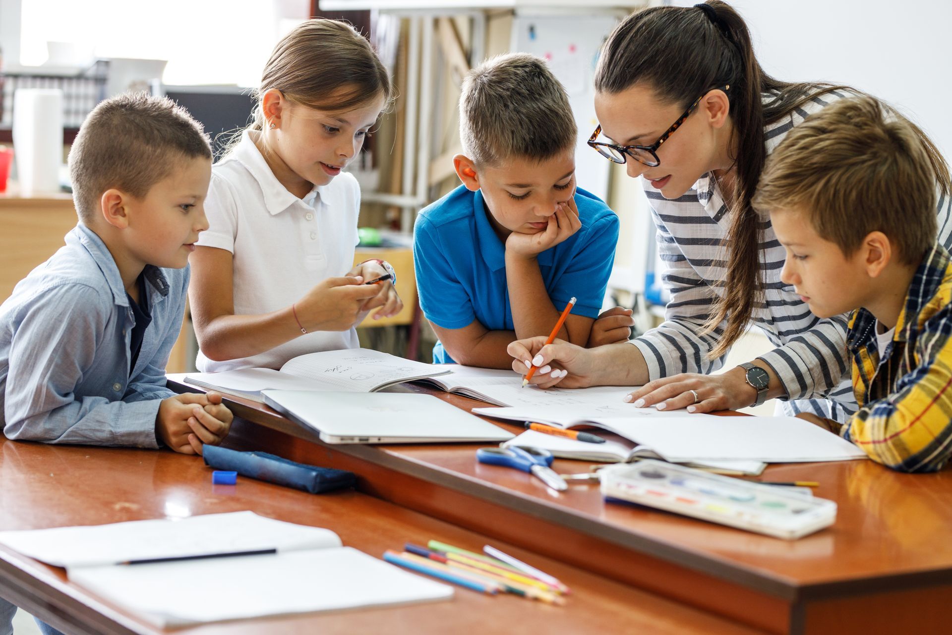 A woman and four children gathered around a desk with the woman showing the children something on a piece of paper that is laying on the desk.