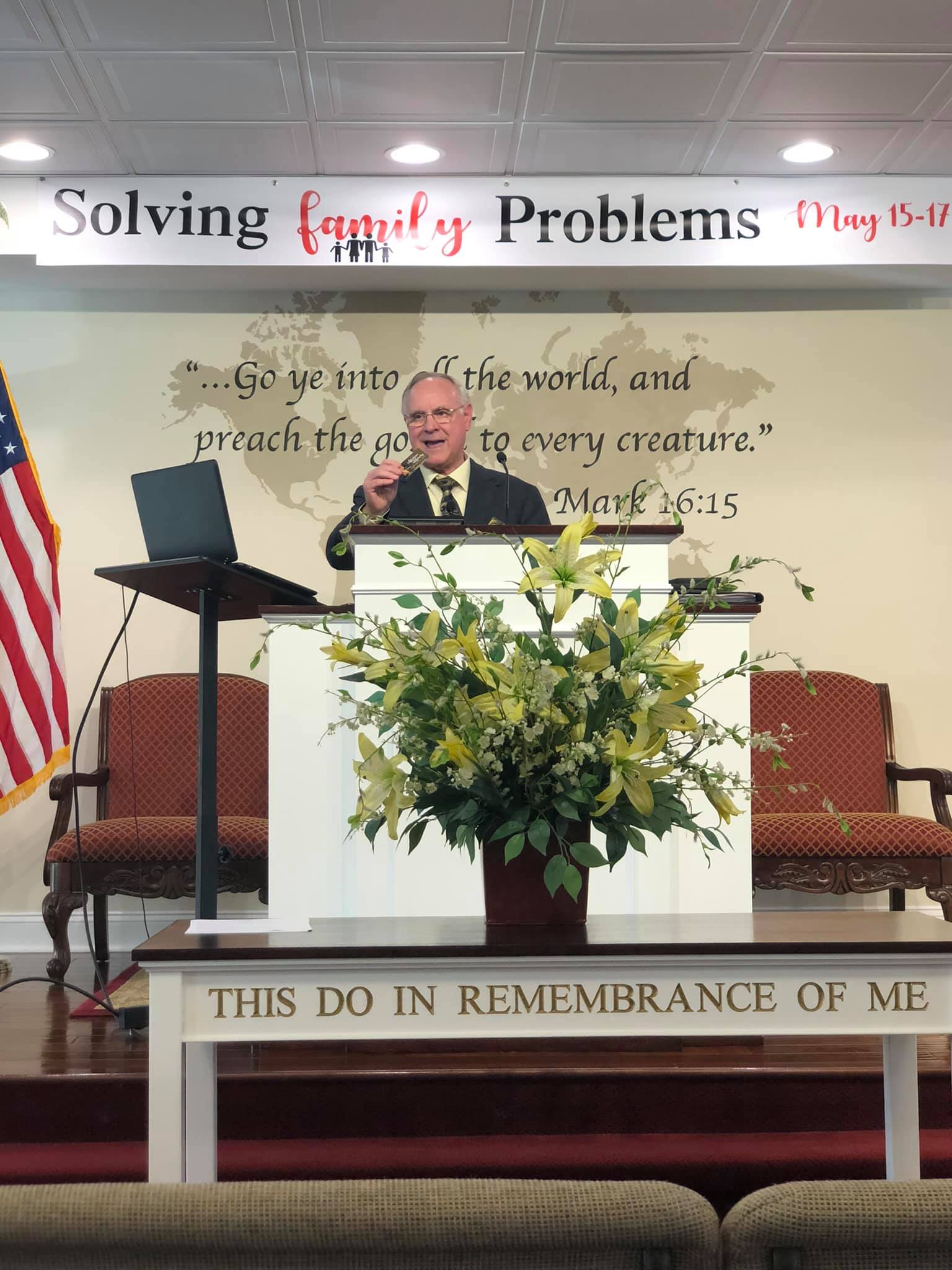 Speaker at church lectern beside floral arrangement and American flag on stage, under “Solving Family Problems” banner