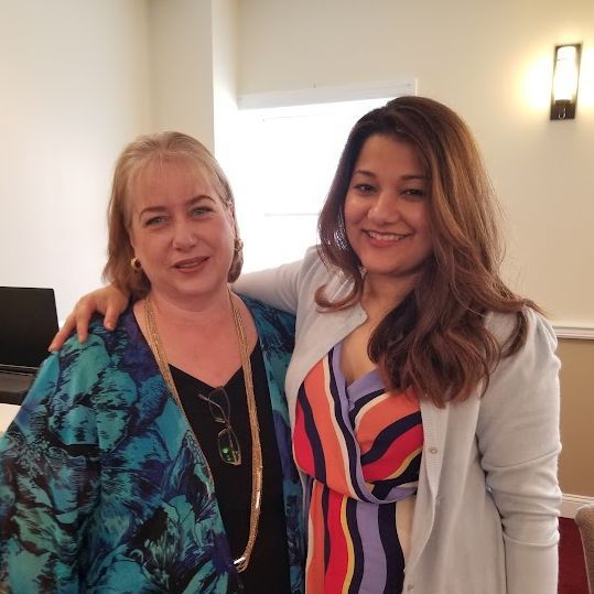 Two women smiling and posing indoors, one arm around the other, wearing colorful outfits.