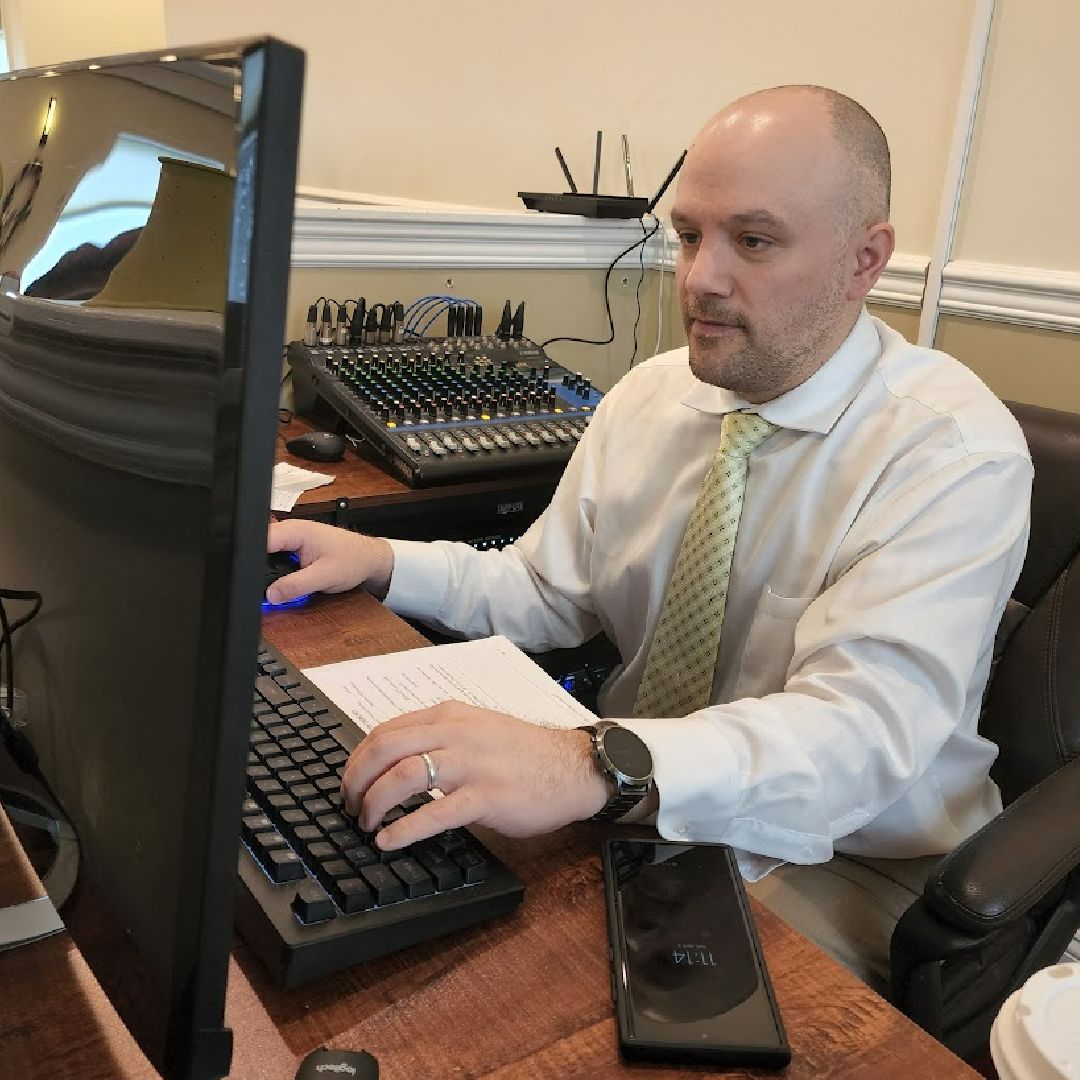 Man at a desk typing on a keyboard beside a computer monitor and audio mixer in an office setting